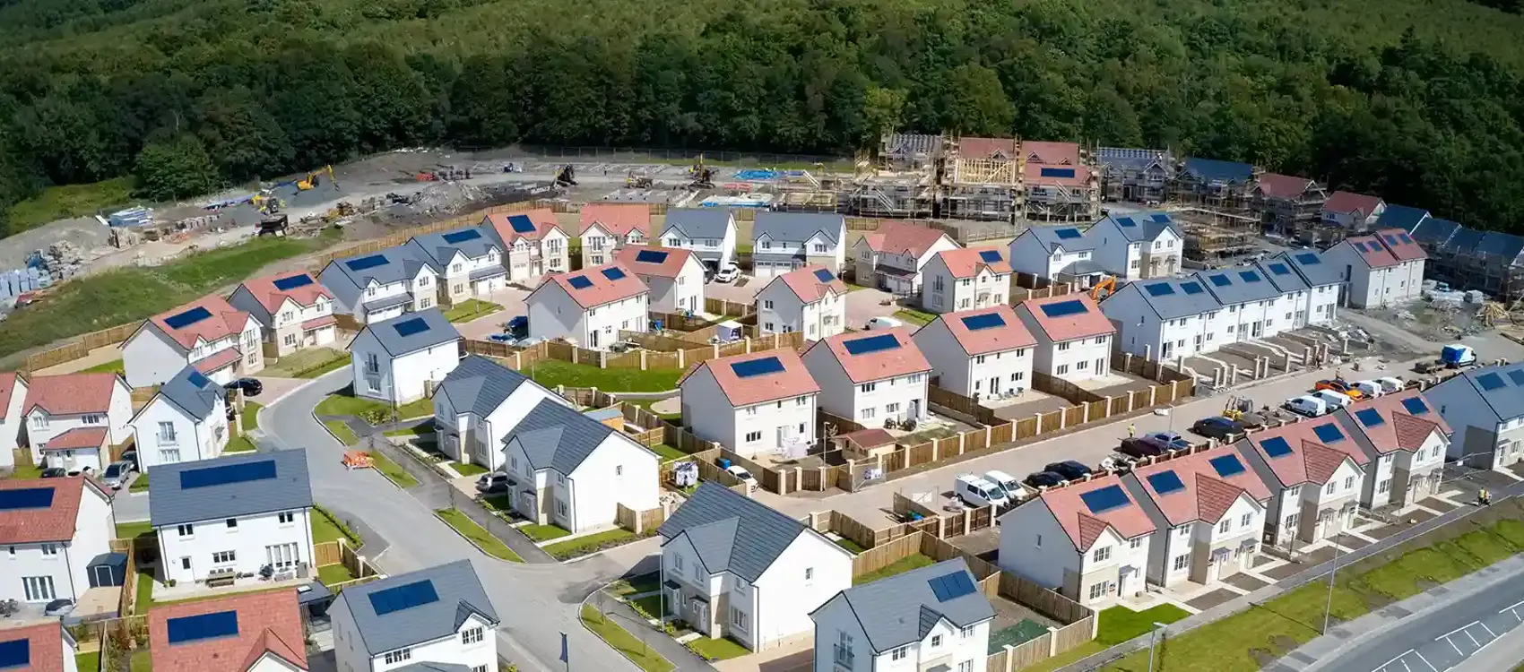 A wide aerial view of a modern residential neighbourhood with solar-panelled homes, surrounded by trees and ongoing construction in the background.