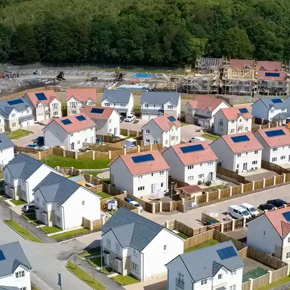 A wide aerial view of a modern residential neighbourhood with solar-panelled homes, surrounded by trees and ongoing construction in the background.