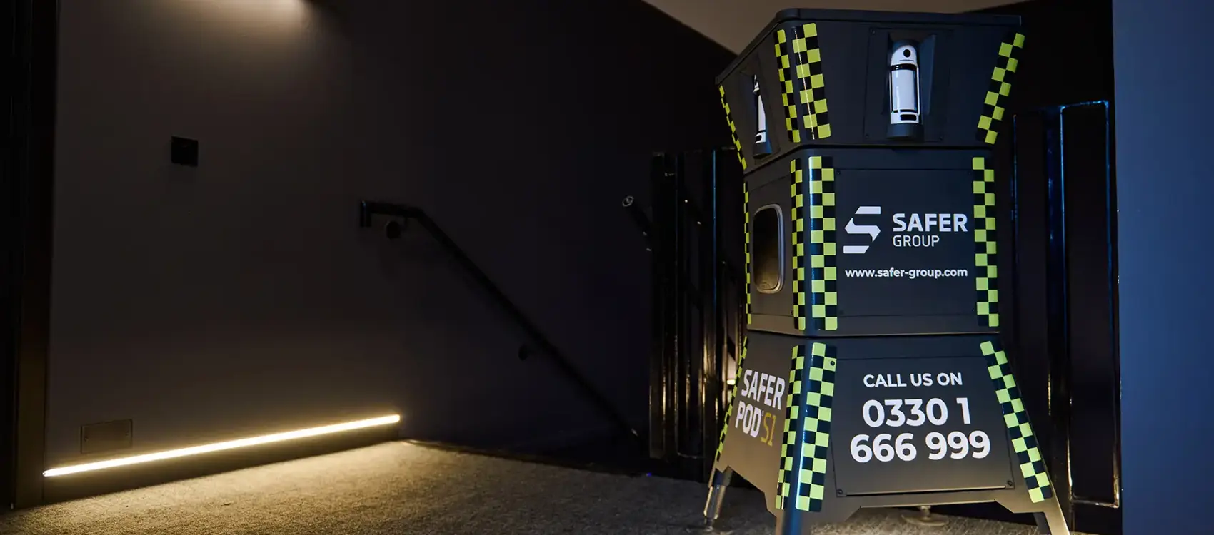 A black and yellow Safer Group Safer POD S1 stands at the top of a staircase, illuminated by soft lighting in a modern space.