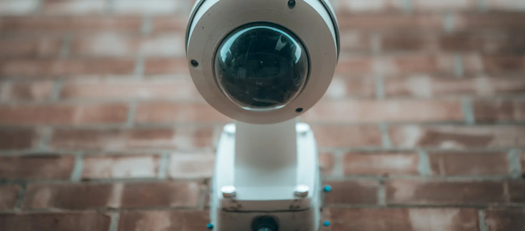 A close-up of a white CCTV security camera mounted on a brick wall, with a steel pipe connecting it to the building.