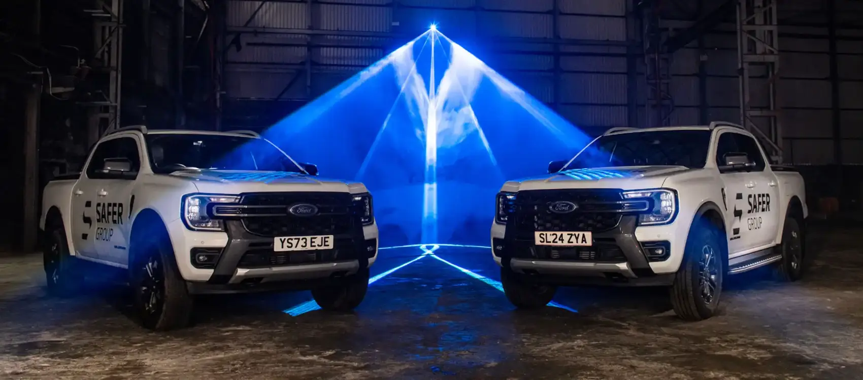 Two white Ford pickup trucks with "Safer Group" branding under dynamic blue lighting in an industrial setting.