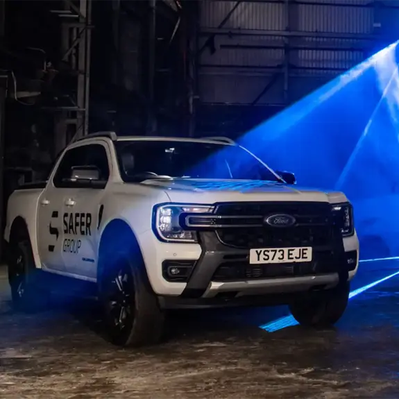 Two white Ford pickup trucks with "Safer Group" branding under dynamic blue lighting in an industrial setting.