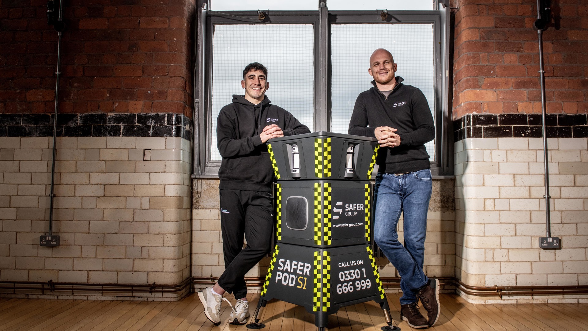 MMA fighter Reece McEwan and Group chief executive Ryan Clark stand leaning on either side of a Safer Pod S1, in a rustic brick-walled room in front of large windows.