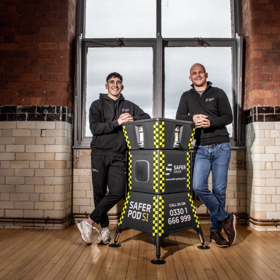 MMA fighter Reece McEwan and Group chief executive Ryan Clark stand leaning on either side of a Safer Pod S1, in a rustic brick-walled room in front of large windows.