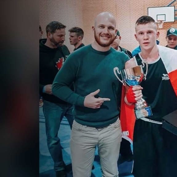 Ryan Clark joins a sponsored athlete celebrating a sports victory, with the athlete holding a trophy while draped in a flag and surrounded by others in a gym hall.