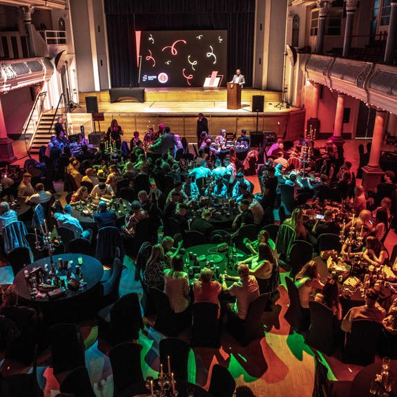 A formal gathering in a grand hall with round tables filled with people under colourful lights. A stage with a speaker and large screen is in the background.