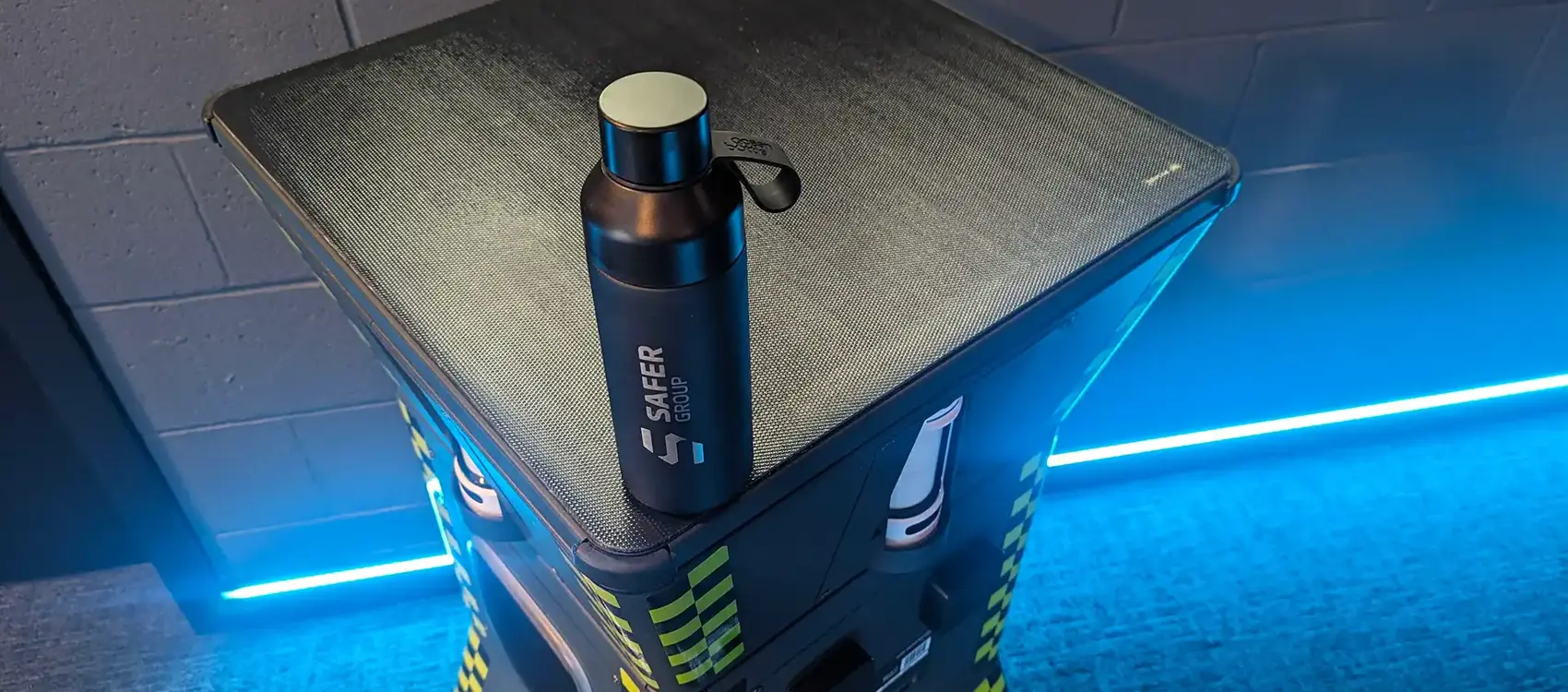 A sleek black water bottle with the "Safer Group" logo rests on a textured solar panel surface with blue LED lighting in the background.