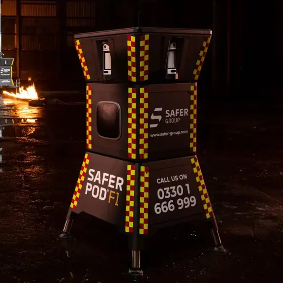 Two black safer pod F1s with yellow and red reflective patterns stand in a dimly lit space, flames flicker in the background alongside a Safer POD F7.