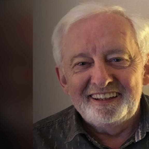 A man with grey hair and trimmed beard wears a patterned shirt, seated in a softly lit room. The background is slightly blurred.