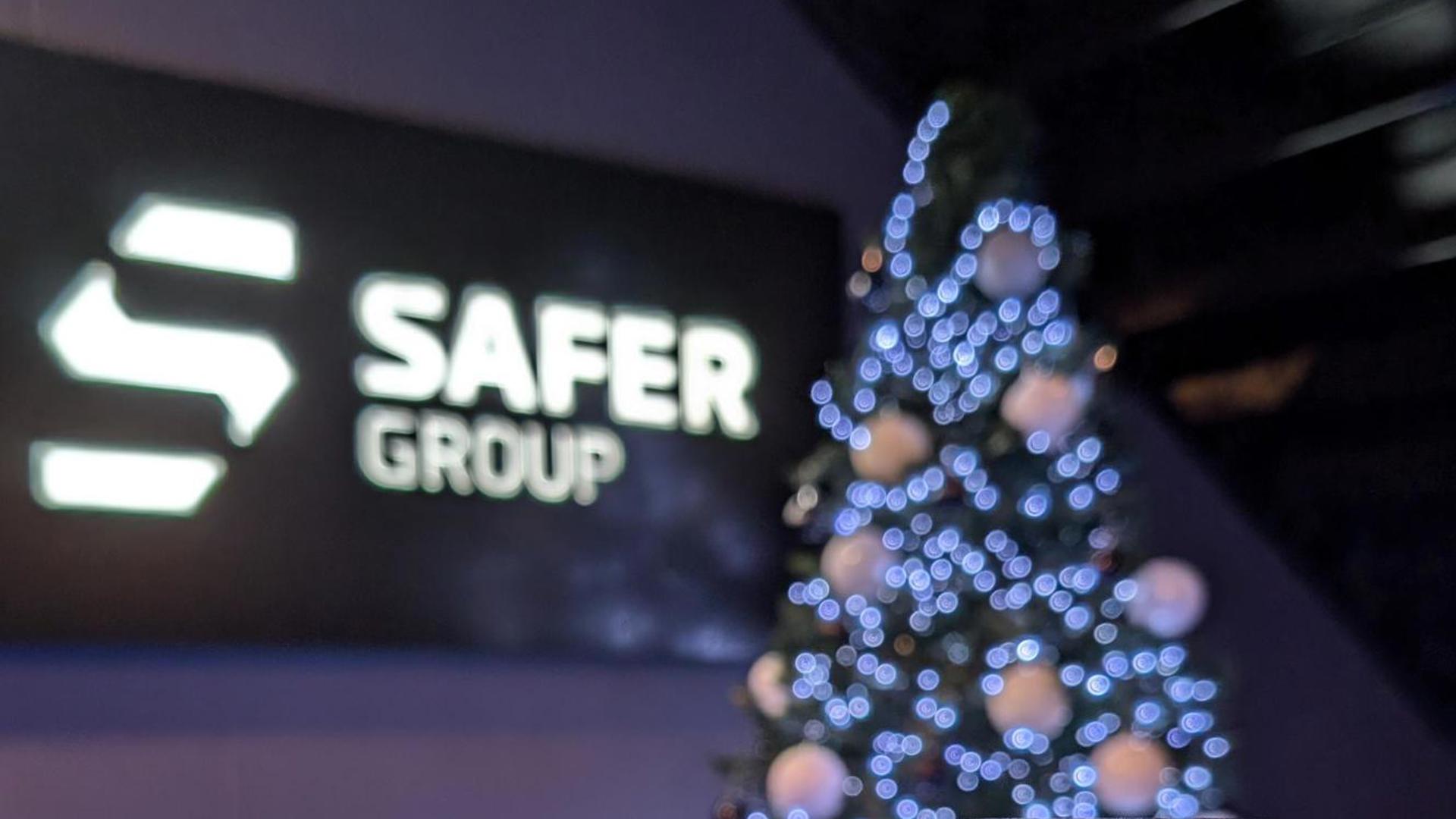 A small toy of a Safer Pod S1 device stands in the foreground, with a Safer Group logo backdrop and a decorated Christmas tree adorned with white lights.