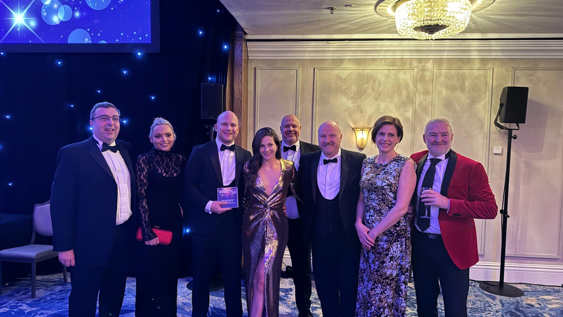 A group of elegantly dressed individuals posing together at a glamorous event, with soft lighting and a starry backdrop.