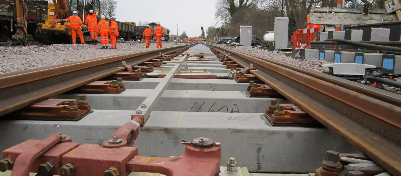Workers in high-visibility orange suits conduct maintenance on railway tracks, with machinery and equipment visible in the background.