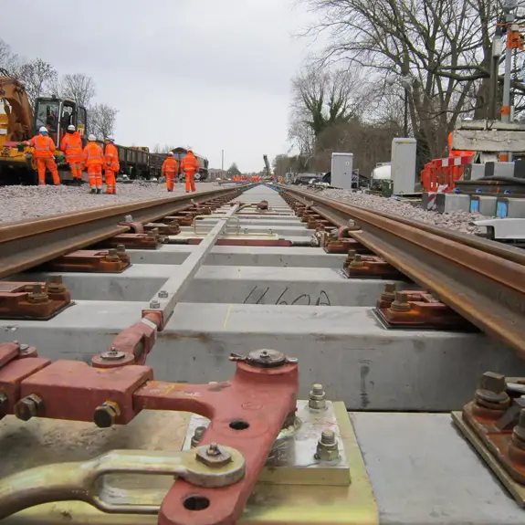 Workers in high-visibility orange suits conduct maintenance on railway tracks, with machinery and equipment visible in the background.