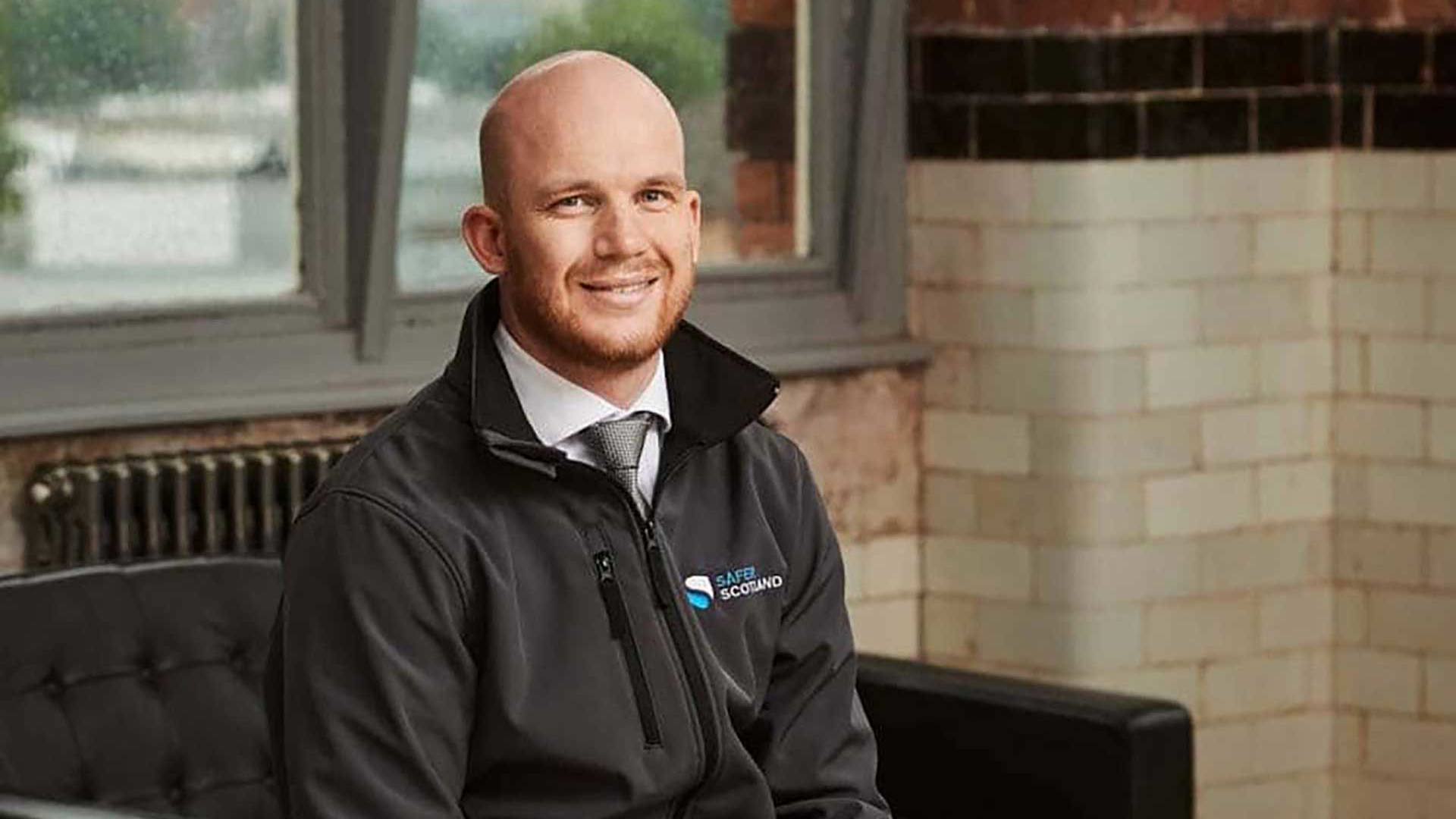 A smiling man in a black jacket with a safer scotland logo on it sits on a black couch in a brick-walled room with large windows.