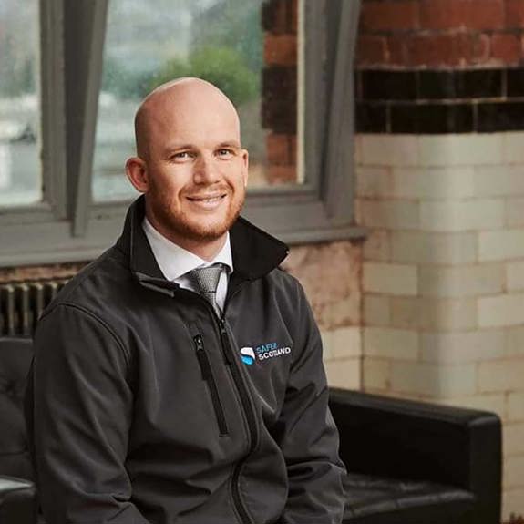 A smiling man in a black jacket with a safer scotland logo on it sits on a black couch in a brick-walled room with large windows.