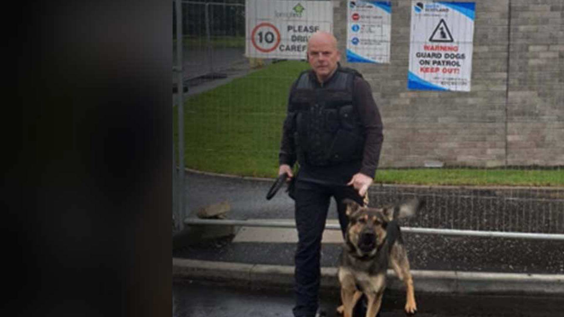 A security officer in tactical gear stands with a German Shepherd outside a gated area. Safer Scotland warning signs are visible nearby.