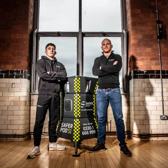 MMA fighter Reece McEwan and Group chief executive Ryan Clark stand confidently flanking a Safer Group S1 pod in a brightly lit, industrial-style interior with large windows.