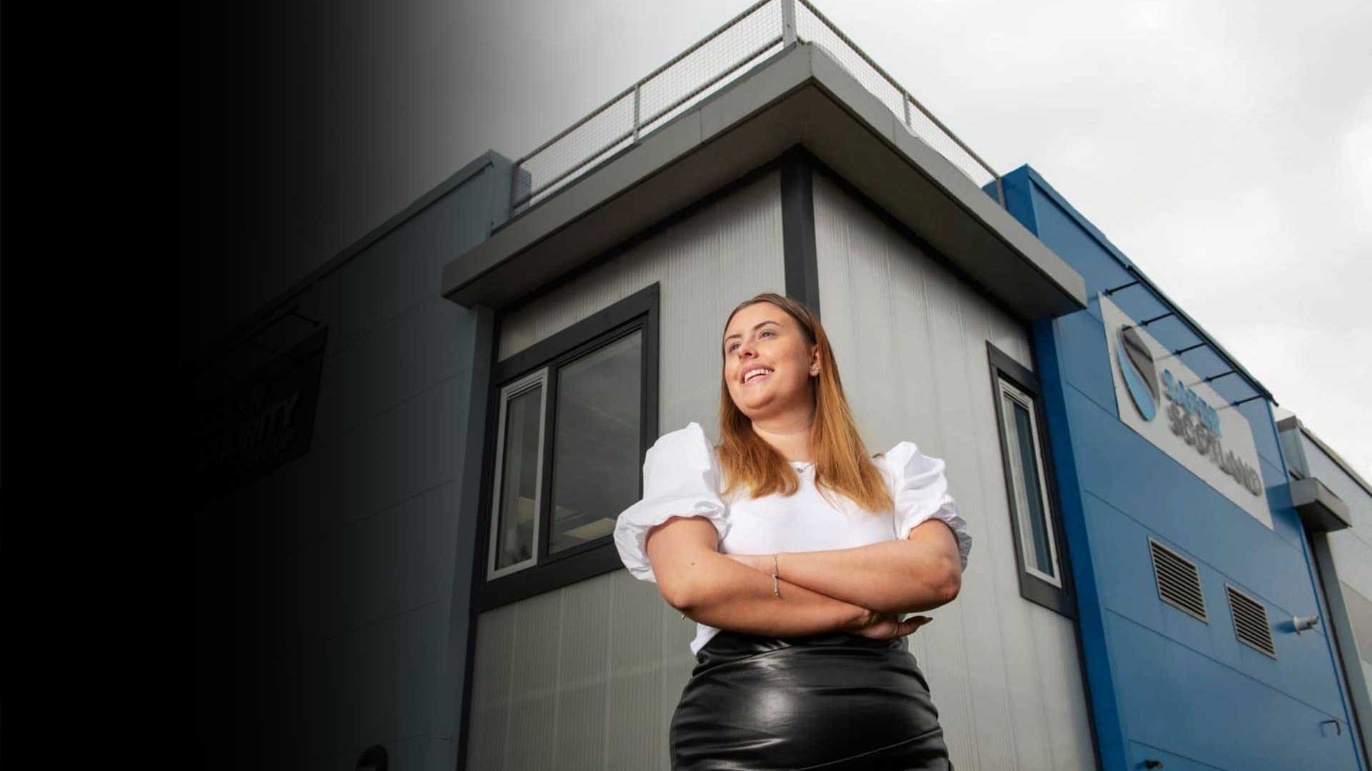 A confident woman stands with arms crossed in front of a modern blue and gray building, with a cloudy sky above.