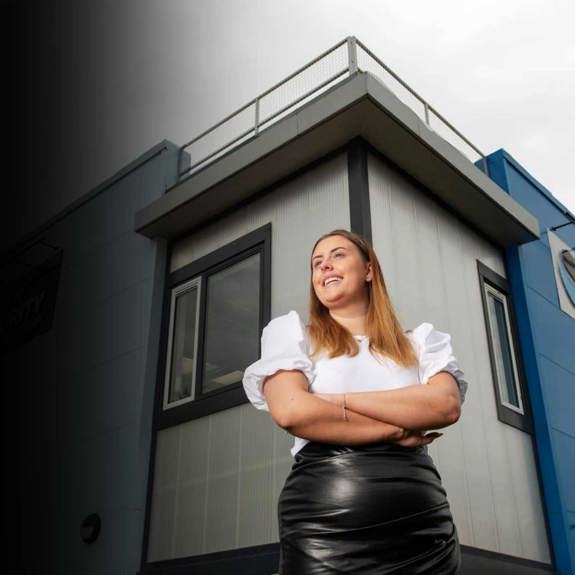 A confident woman stands with arms crossed in front of a modern blue and gray building, with a cloudy sky above.