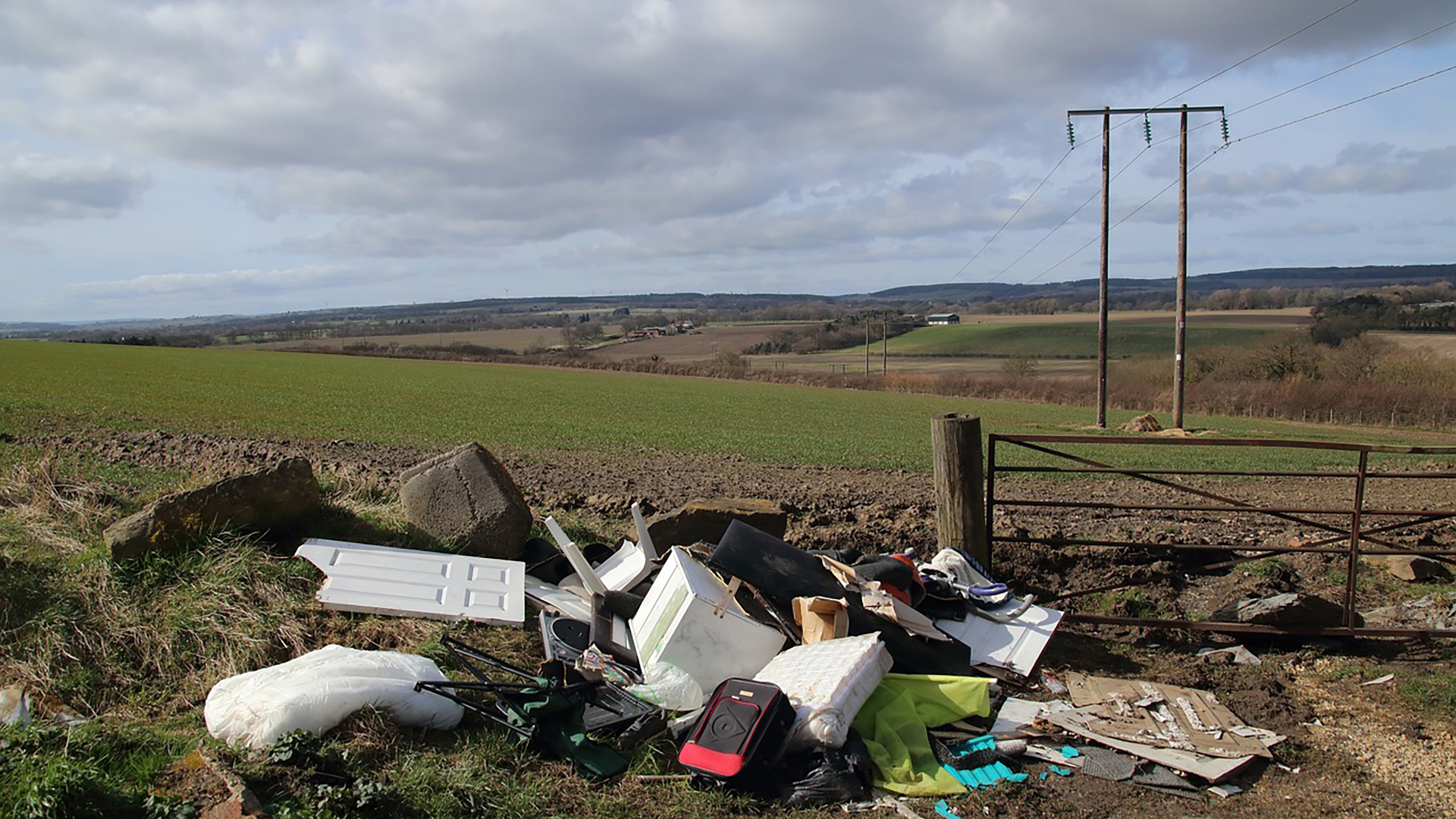 A pile of discarded household items and rubbish near an open field, with rolling hills and cloudy sky in the background.