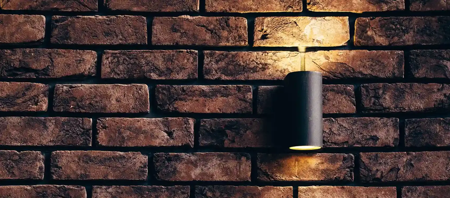A textured, warm-toned brick wall illuminated by a minimalist black wall lamp, creating a cozy atmosphere.